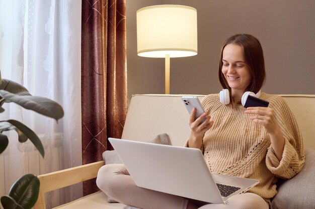 Kaukasisch meisje dat thuis online winkelt Jonge vrouw die op de bank zit met creditcard smartphone en laptop bestelt op mobiele telefoon app betaalt op internet