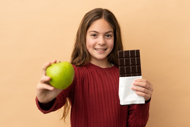 Kaukasisch meisje geïsoleerd op een beige achtergrond met een chocoladetablet in de ene hand en een appel in de andere the