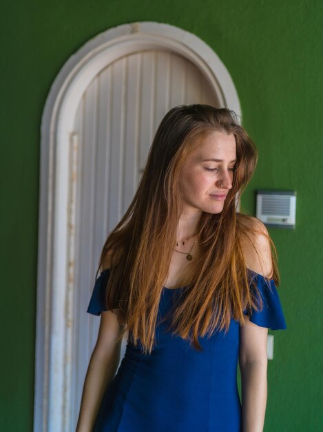 Kaukasisch meisje in een blauwe jurk naast een witte deur van een verticale foto van een groen huis