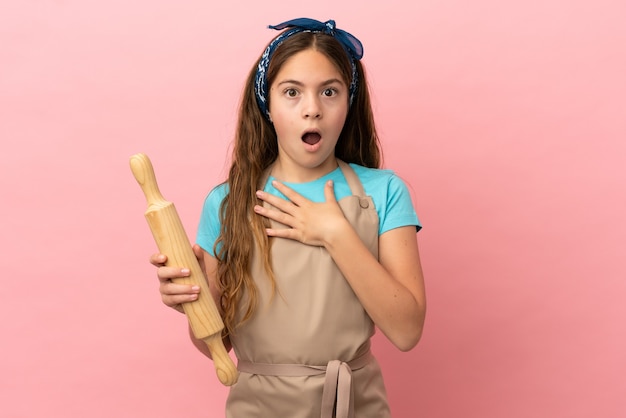 Kaukasisch meisje met een deegroller geïsoleerd op roze achtergrond verrast en geschokt terwijl ze naar rechts kijkt