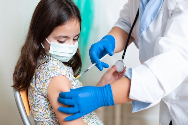 Kaukasisch meisje met gezichtsmasker dat toekijkt hoe ze wordt gevaccineerd door een medisch werker. Kind vaccinatie concept.