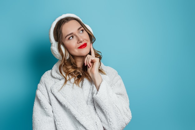 Kaukasisch meisje met rode lippenstift gekleed in een witte winterjas van imitatiebont en een koptelefoon van bont is attent en kijkt naar de zijkant naar kopie ruimte voor tekst geïsoleerd op blauwe muur