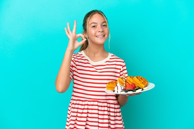 Kaukasisch meisje met wafels geïsoleerd op een blauwe achtergrond met ok teken met vingers