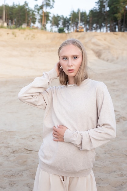 Kaukasisch mooi jong meisje met lang haar en naakt make-up in beige kleding vormt buiten op zand voor bos