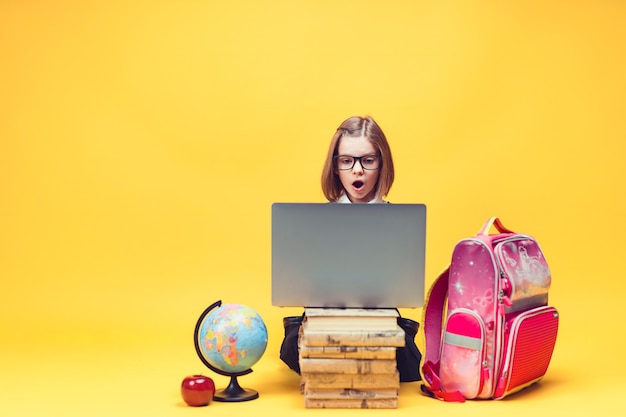 Kaukasisch schoolmeisje in shock zittend achter een stapel boeken kijkend naar het onderwijs van laptops voor kinderen