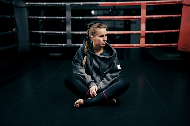 Kaukasisch sterk boksermeisje dat in ring in sweatshirt en met verband op voeten zit en weg kijkt.