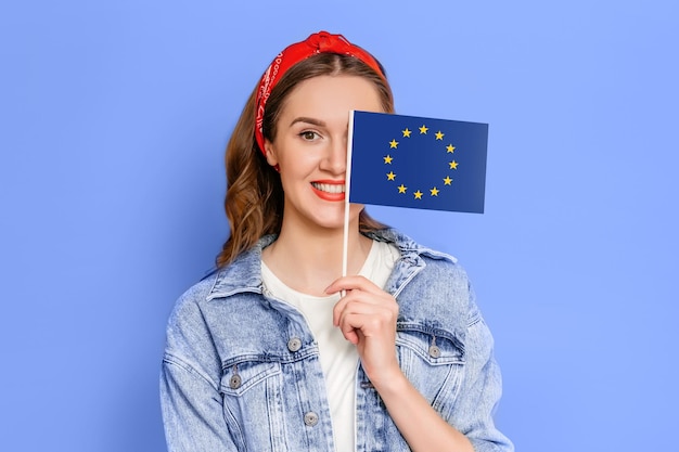 Foto kaukasisch studentenmeisje die kleine vlag met het embleem van de europese unie houden dat over blauwe achtergrond wordt geïsoleerd