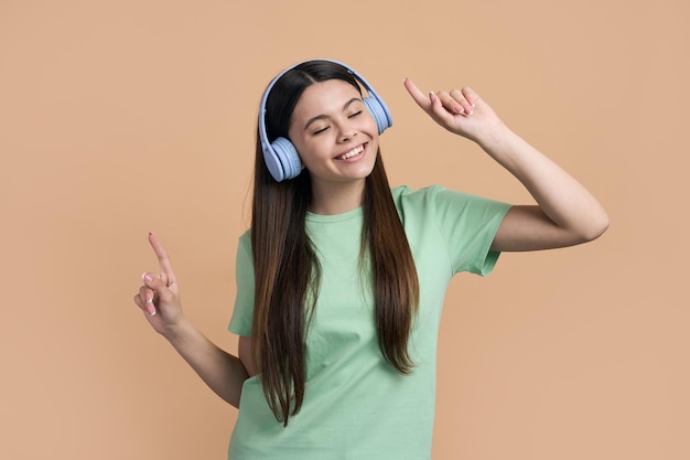 Kaukasisch vrolijk gelukkig lachend tienermeisje dansen luisteren naar muziek op blauwe draadloze hoofdtelefoons Mooi adolescent kind veel plezier punten vingers terwijl dansen over geïsoleerde beige kleur achtergrond