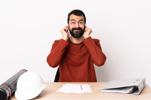 Kaukasische architectenmens met baard in een gefrustreerde tafel en behandelende oren.