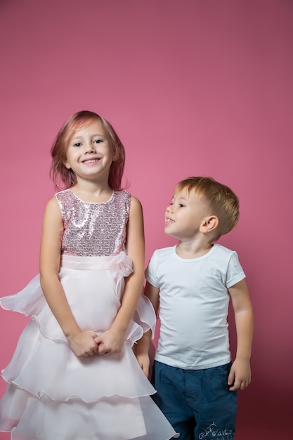 Kaukasische broer en zus, knuffelen op camera op roze achtergrond studio opname. Familiebanden, vriendschap, gelukkig jeugdconcept.