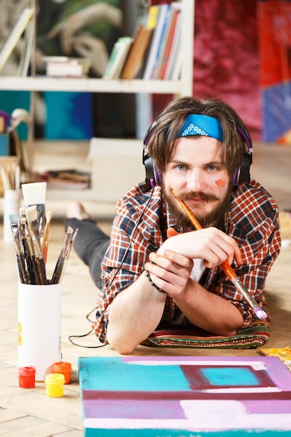 Kaukasische donkerharige bebaarde hipster mannelijke artiest in koptelefoon en blauwe bandana ligt op de vloer en houdt borstel vast in zijn hedendaagse studio met veel schilderijen