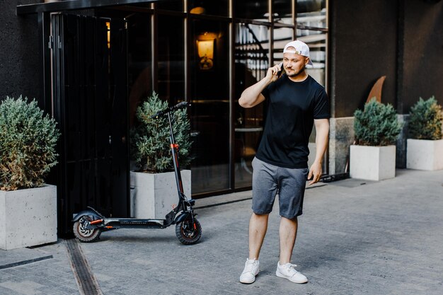 Kaukasische jonge knappe man die op straat op een elektrische scooter staat en op mobiele telefoon praat Stijlvolle man die buitenshuis op mobiele telefoon spreekt