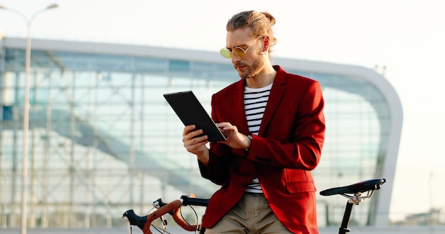 Kaukasische jonge knappe man die zich bij fiets bevindt en tabletapparaat gebruikt. stijlvolle man in rode jas en zonnebril, leunend op de fiets en tikken of scrollen op de computer van de gadget op modern glazen gebouw.