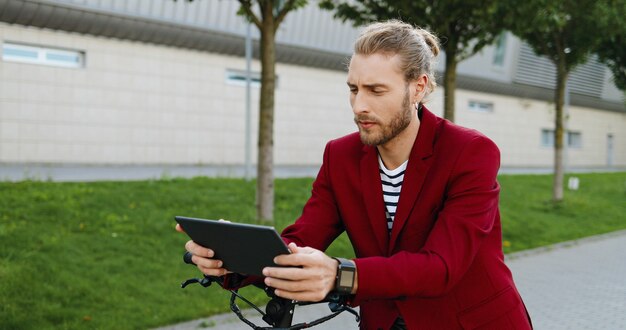 Kaukasische jonge knappe man in rode jas staande op straat op elektrische scooter en kijken naar video op tablet-apparaat. Stijlvolle man tikken en scrollen op gadgetcomputer. Sms'en op sociale media.