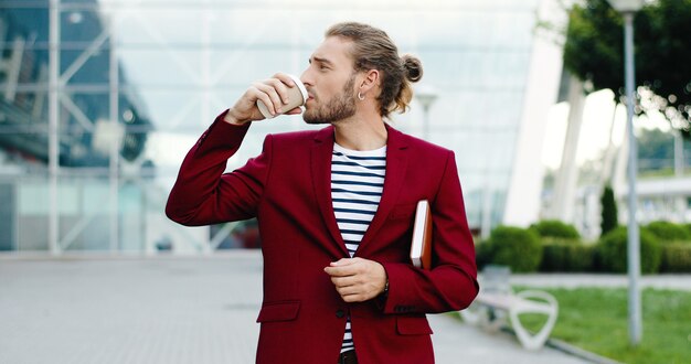 Kaukasische jonge knappe stijlvolle zakenman buiten wandelen in de stad met planner en nippend drankje. Zakencentrum modern gebouw op de achtergrond. Mannetje wandelen en koffie drinken om mee te nemen.