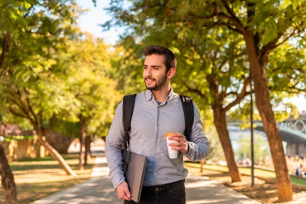 Kaukasische jonge student en werkman die een afhaalkoffie drinkt en op een zonnige dag zijn laptop in een prachtig park draagt.