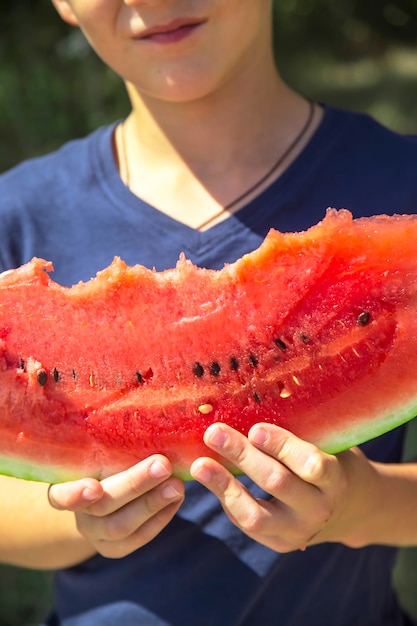 Kaukasische jongen die en een grote gebeten plak van rode rijpe watermeloen houdt eet