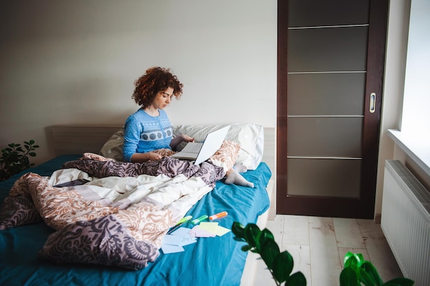 Kaukasische krullende vrouw doet online lessen vanuit bed met behulp van laptop in een blauwe pyjama quarantaineleven...