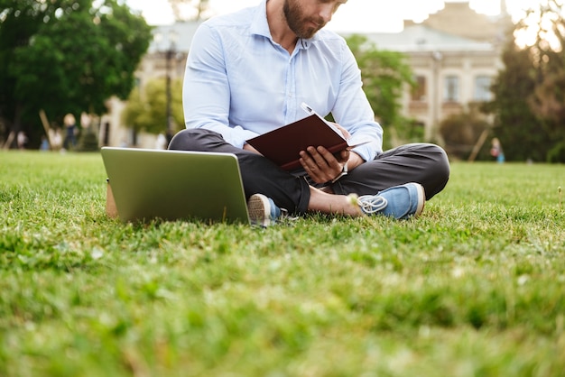 Kaukasische man met wit overhemd, notities in notitieblok opschrijven terwijl zittend op gras in park met gekruiste benen en bezig met laptop