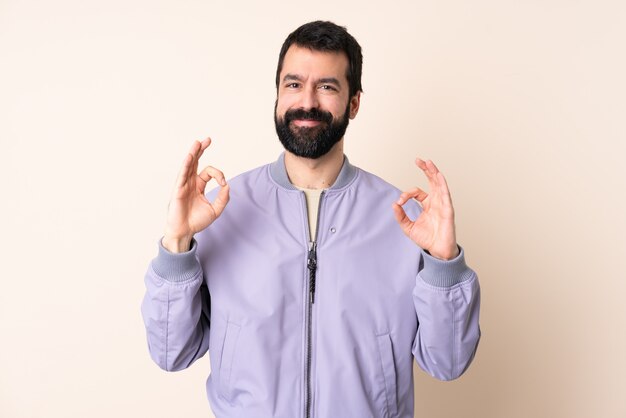 Kaukasische mens met baard die een jasje over geïsoleerde muur draagt die ok teken met twee handen toont