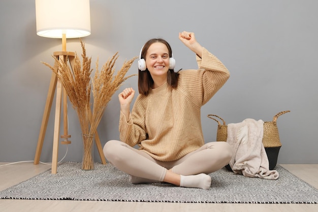 Kaukasische mooie vrouw die thuis glimlacht in koptelefoon, beige trui draagt, naar muziek luistert, moderne technologie gebruikt voor vrije tijd, gelukkig danst met opgeheven armen.