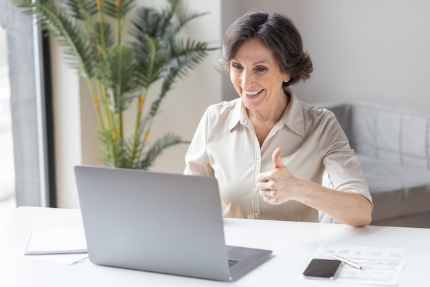 Kaukasische oude zakenvrouw zwaait met de hand, begroet de deelnemers aan de videoconferentie, gebruikte laptopcomputer om online te communiceren op kantoor of thuis. Videogesprek, concept voor werken op afstand