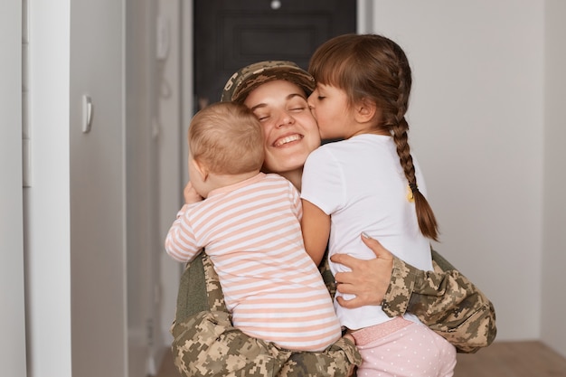 Kaukasische patriottische jonge volwassen vrouwelijke soldaat die camouflage-uniform draagt, terugkeert, kinderen die moeder kussen na haar thuiskomst uit oorlog of leger, gelukkige ontmoeting.