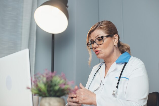 Kaukasische senior medic spreekt online met de patiënt die een laptop gebruikt en een bril draagt terwijl hij voor de camera spreekt