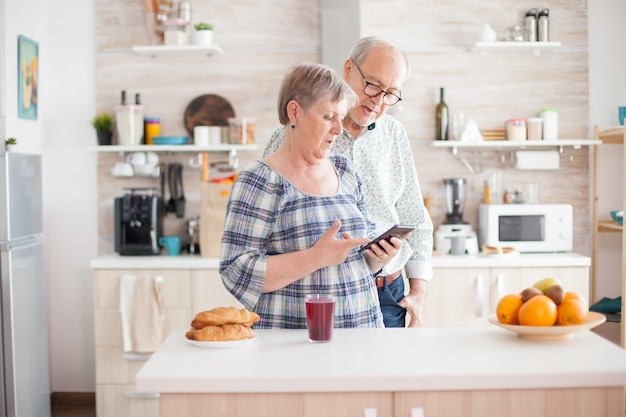 Kaukasische senior vrouw en echtgenoot in de keuken tijdens het ontbijt met behulp van smartphonetechnologie en internetverbinding. Vrije tijd gepensioneerde vrouw en man die lacht in woonplaats, ontspannen.