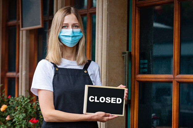 Kaukasische serveerster met een medisch masker houdt een bordje vast dat de coronavirus-pandemie is gesloten.