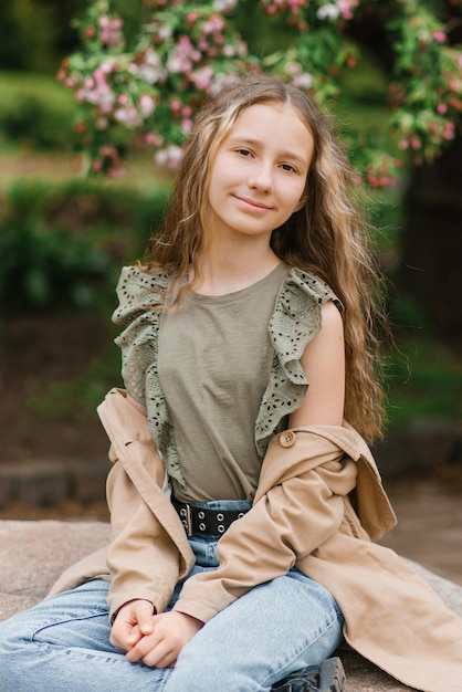 Foto kaukasische tienermeisje met lang blond haar in een trenchcoat zit op een rots in het park