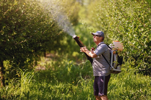 Kaukasische volwassen boer in werkkleding, hoed en met moderne pesticidespuitmachine op ruggen die insecten in boomgaard sproeien.