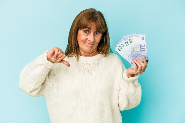 Kaukasische vrouw van middelbare leeftijd met bankbiljetten geïsoleerd op blauwe achtergrond met een afkeer gebaar, duim omlaag. Onenigheid begrip.