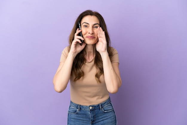 Kaukasische vrouw van middelbare leeftijd met behulp van mobiele telefoon geïsoleerd op paarse achtergrond schreeuwen met wijd open mond