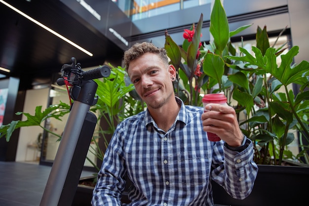 Kaukasische zakenman gekleed in een geruit overhemd drinkt koffie zittend op een bankje met zijn elektrische scooter. glimlachende duizendjarige jongen wijst met zijn vinger naar de camera - duurzaam mobiliteitsconcept