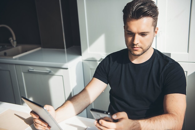 Kaukasische zakenman werkt op afstand thuis met behulp van een telefoon en digitale tablet poseren in de keuken