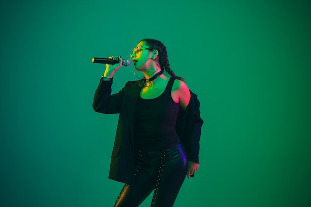 Kaukasische zangeres portret geïsoleerd op groene muur in neonlicht. Mooi vrouwelijk model in zwarte slijtage met microfoon. Concept van menselijke emoties, gezichtsuitdrukking, advertentie, muziek, kunst.