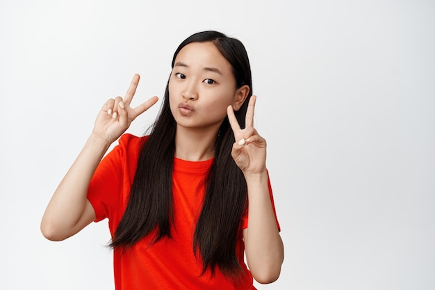 Азиатская девушка Kawaii показывает целующиеся губы и жест мирного v-знака, позирует и выглядит мило на белом, стоя в красной футболке на белом