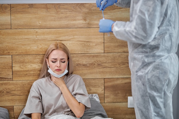 Keelpijn in het coronavirusseizoen van de griep. vrouw raakt haar nek aan en voelt pijn terwijl ze thuis in de slaapkamer zit terwijl de arts zich voorbereidt op een medische test, die lijdt aan covid-19-symptomen.