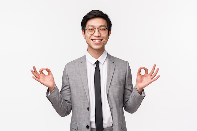 Keep calm and stay healthy. Handsome cheerful smiling asian male entrepreneur, office worker staying calm, holding hands in zen gesture, relaxing, meditating, on a white wall
