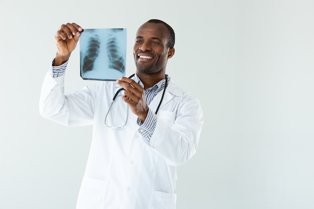 Следить. Веселый афроамериканский мужчина держит в руках рентгеновский снимок легких во время работы