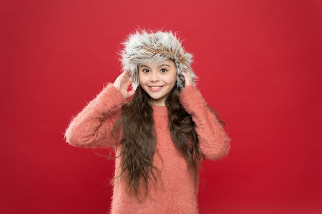 Keep head in warmth. Feeling warm. Child long hair soft hat. Winter fashion concept. Warm hat for cold winter weather. Kid girl red background. Soft furry accessory. Winter season. Shopping concept.