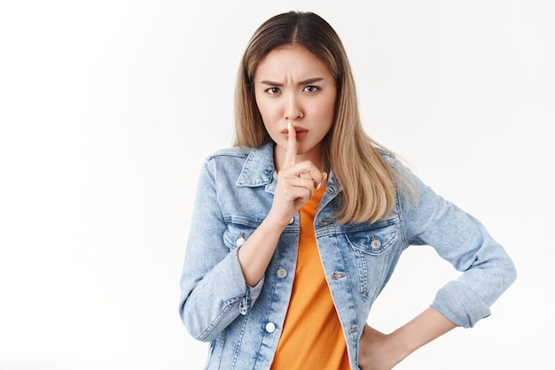 Keep mouth shut please Seriouslooking intense bothered blond asian woman shush hold index finger mouth prohibiting talk loud frowning displeased say shhh angry standing white background