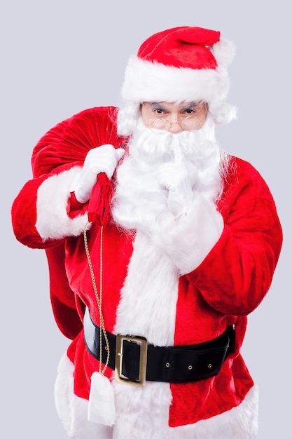 Keep my secret! Portrait of traditional Santa Claus holding his finger on lips while carrying sack with presents and standing against grey background