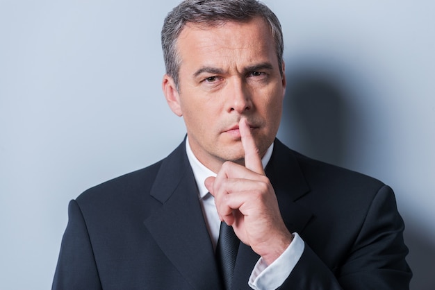 Keep my secret!  Serious mature man in formalwear holding finger on lips and looking at camera while standing against grey background
