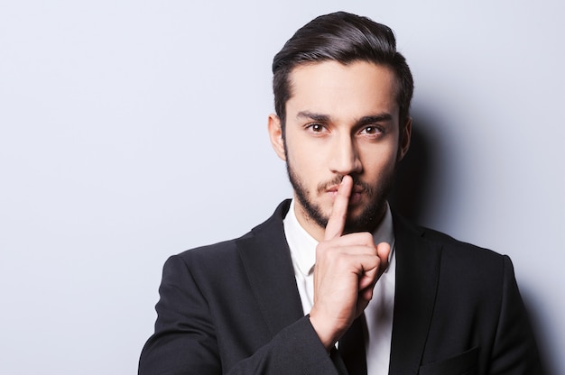 Keep my secret!  Serious young man in formalwear holding finger on lips and looking at camera while standing against grey background