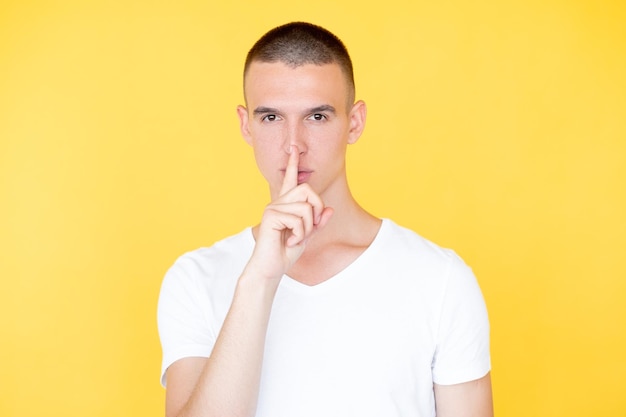 Keep secret Silent man Mute expression Serious hipster guy in casual white tshirt holding shush finger on mouth isolated yellow