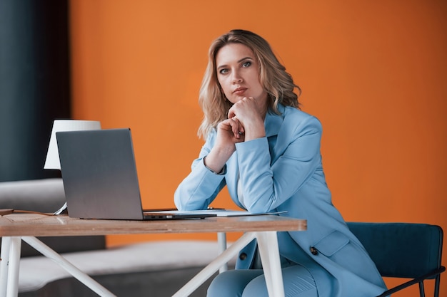 Keep talking, I listen to you. Businesswoman with curly blonde hair indoors in room with orange colored wall and wooden table.