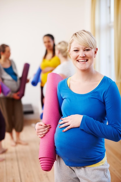 Keeping healthy for my little one A young blonde pregnant woman in a gym holding an exercise mat with a group of women in the background