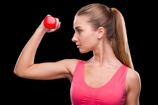 Keeping her body fit. Sporty young woman exercising with dumbbell while standing against black background